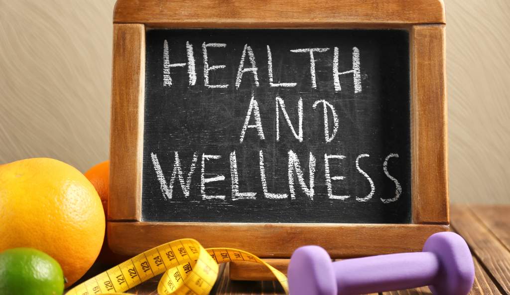 A small chalkboard with 'Health and Wellness' written on it, surrounded by fresh fruits, a measuring tape, and a purple dumbbell, symbolizing a healthy lifestyle