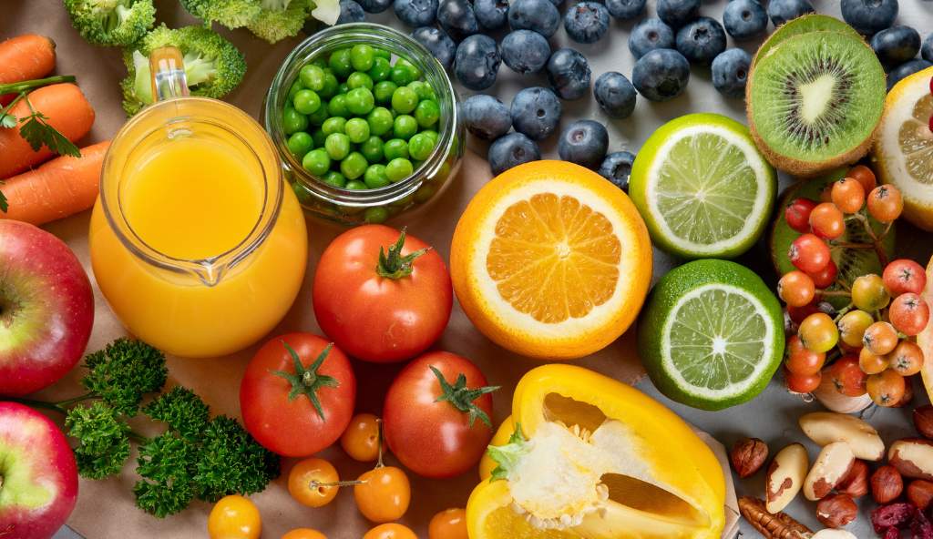 Assortment of fresh fruits and vegetables, including oranges, limes, tomatoes, apples, blueberries, carrots, peas, and a jug of orange juice, symbolizing healthy eating and nutrition