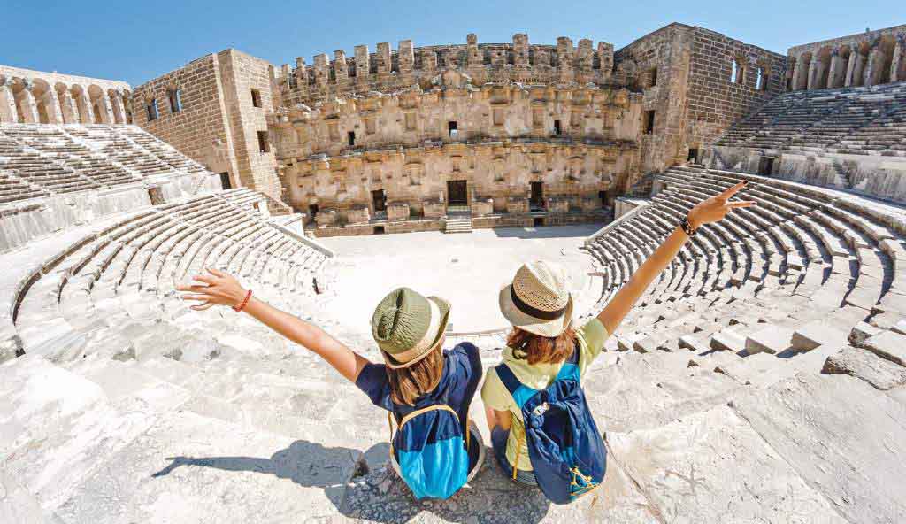 Two travelers wearing straw hats and backpacks sitting on stone steps of an ancient Roman amphitheater, raising their arms in excitement while admiring the historic ruins vacation