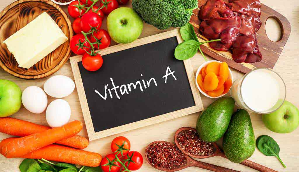 Various foods rich in Vitamin A, including carrots, eggs, tomatoes, butter, green apples, broccoli, avocados, liver, dried apricots, milk, and spinach, arranged around a small chalkboard with 'Vitamin A' written on it.