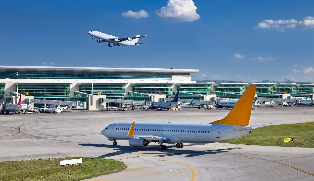 Busy airport terminal with multiple airplanes; one plane taxiing on the runway while another takes off in the background under a clear blue sky flight