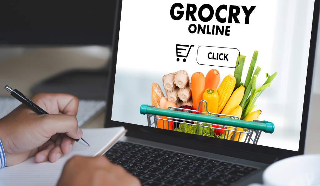 A person taking notes while looking at an online grocery shopping website on a laptop screen, which features a shopping cart filled with fresh vegetables and a 'Click' button grocery