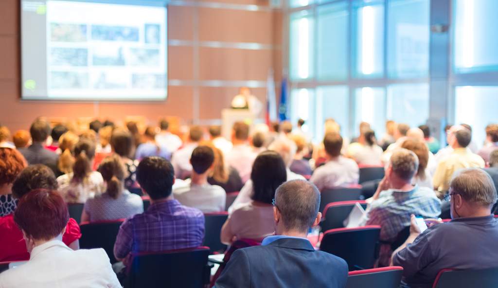 Large audience attending a conference, listening to a speaker giving a presentation in a modern, well-lit conference hall events