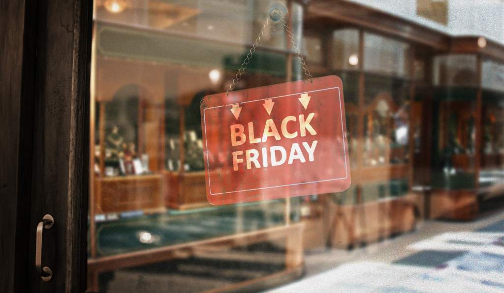 A red sign with white text reading 'BLACK FRIDAY' hangs on a glass storefront, reflecting a shopping mall with warm lighting and wooden display cases in the background