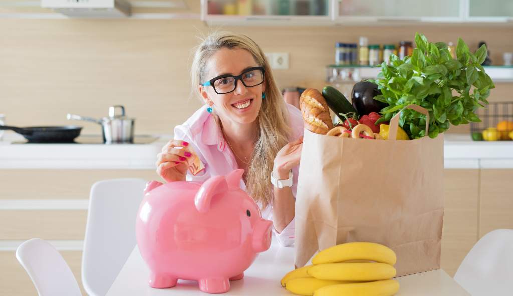 Smiling woman in glasses sitting in a kitchen with a pink piggy bank and a paper bag filled with fresh groceries, symbolizing budgeting and smart shopping bundles