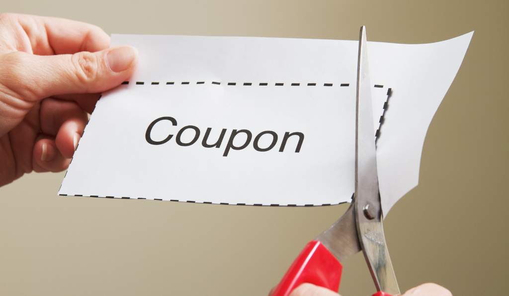 Close-up of hands cutting out a coupon with red-handled scissors, following a dotted outline on white paper printed