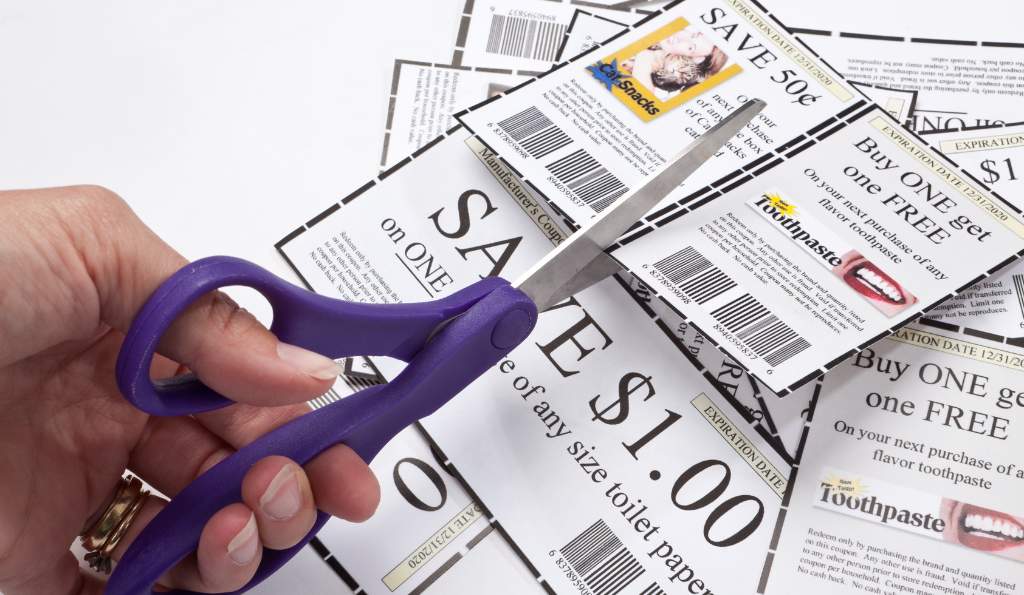 Close-up of a hand using purple scissors to cut out discount coupons offering savings and buy-one-get-one-free deals on various products printed