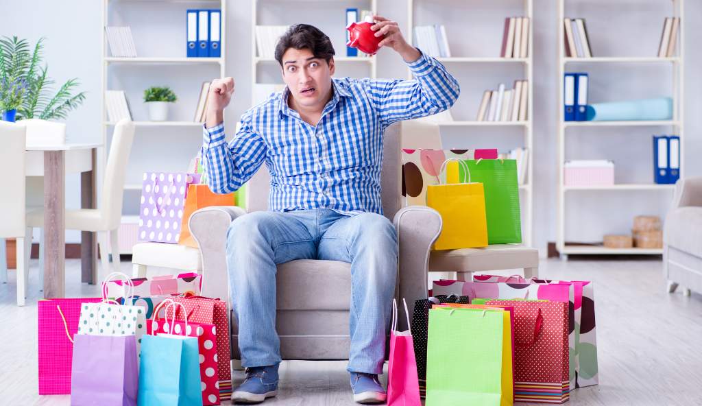 Shocked man sitting in a chair surrounded by colorful shopping bags, holding an empty red piggy bank, depicting overspending and financial stress bundles