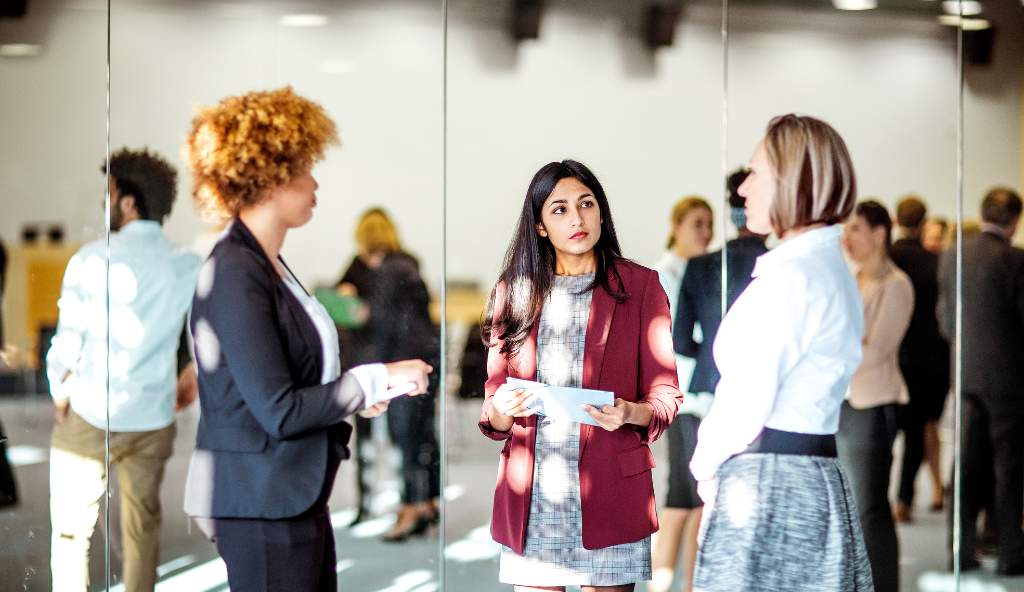 Three professionally dressed women engaged in a discussion at a business networking event, with other attendees conversing in the background events