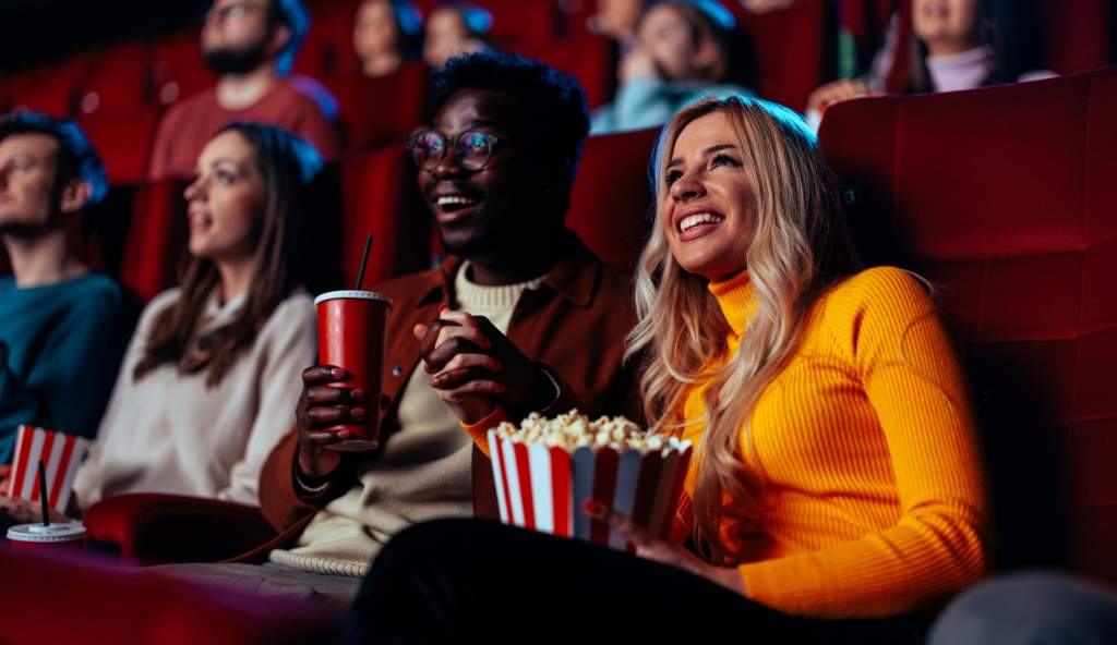 Excited audience in a movie theater, with a smiling couple holding popcorn and drinks while watching a film