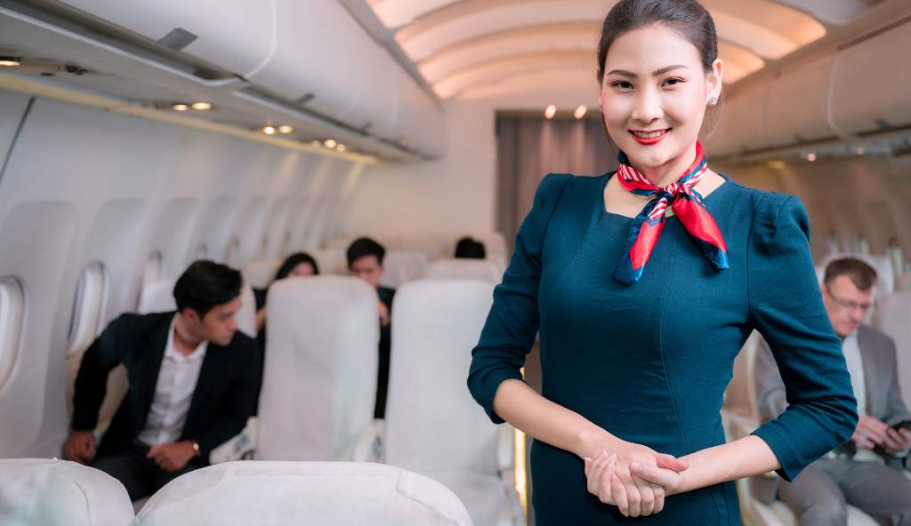 Smiling flight attendant in a navy blue uniform with a red neck scarf standing inside an airplane cabin, with passengers seated in the background flight