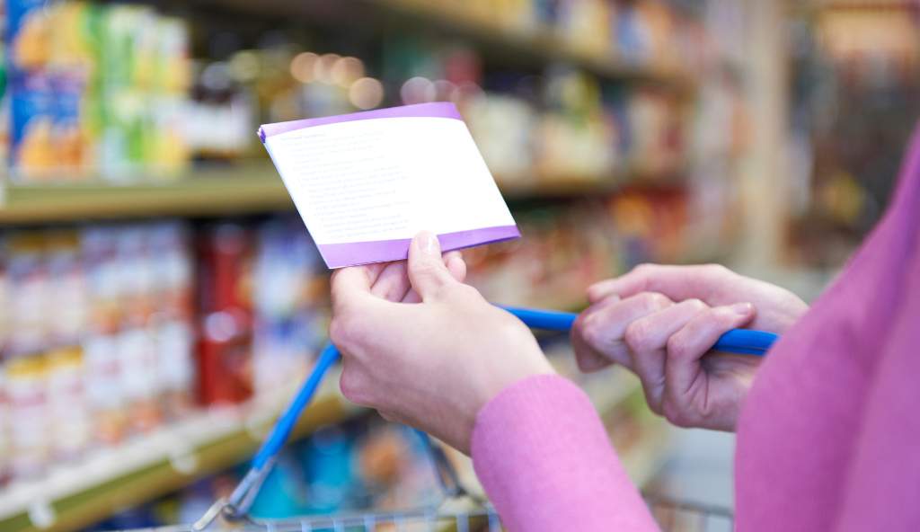 Close-up of a person holding a shopping list while pushing a cart in a grocery store aisle, focusing on budgeting and planned shopping bundles