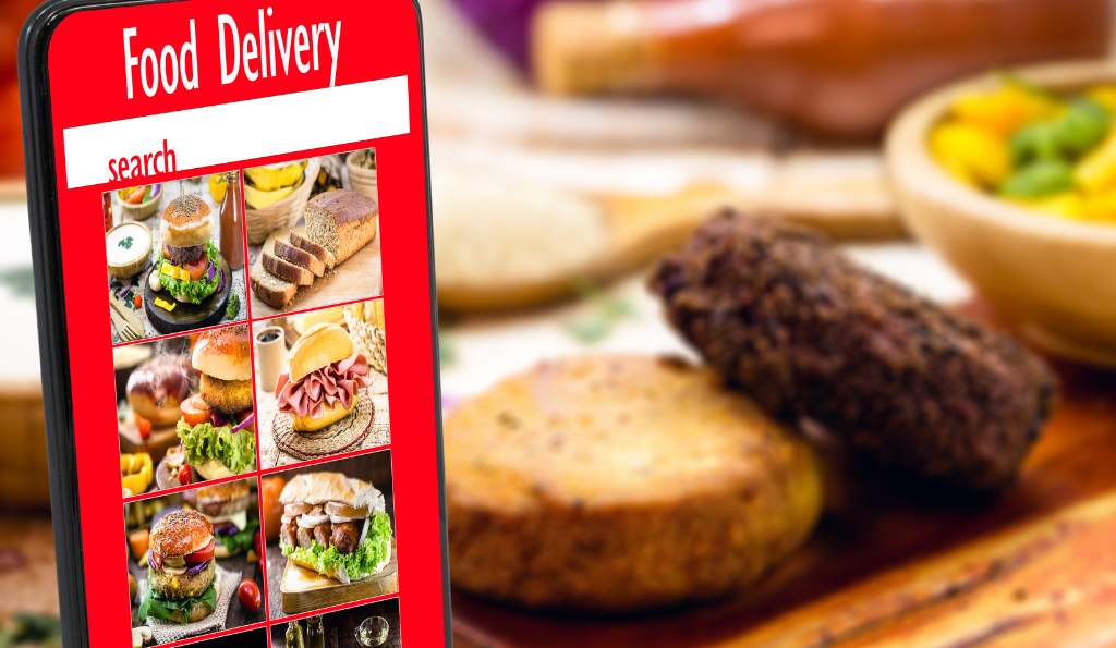 A smartphone screen displaying a food delivery app with various burger and sandwich images, placed in front of a blurred background featuring a wooden table with grilled food and a bowl of vegetables