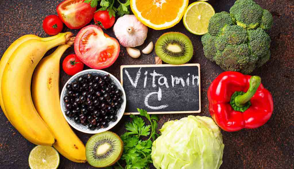 Various fresh fruits and vegetables rich in Vitamin C, including bananas, tomatoes, oranges, lemons, kiwi, broccoli, red bell pepper, black currants, garlic, and cabbage, arranged around a small chalkboard with 'Vitamin C' written on it