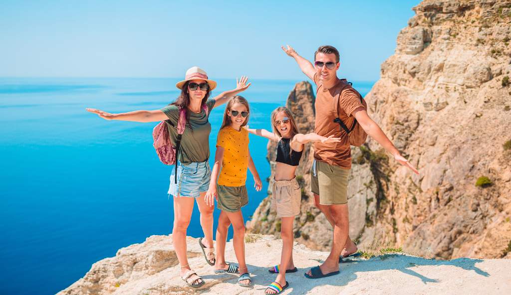 Happy family of four with backpacks posing on a rocky cliff overlooking the blue sea, arms outstretched, enjoying a scenic summer hike vacation