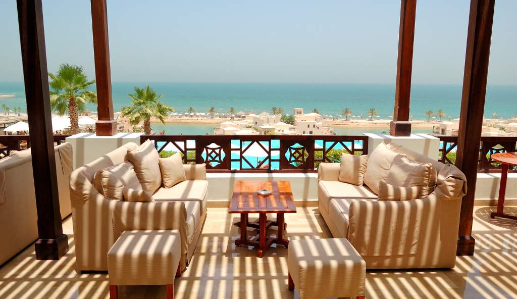 Luxury outdoor lounge with beige sofas and wooden furniture on a terrace overlooking the ocean, a resort, and palm trees hotel