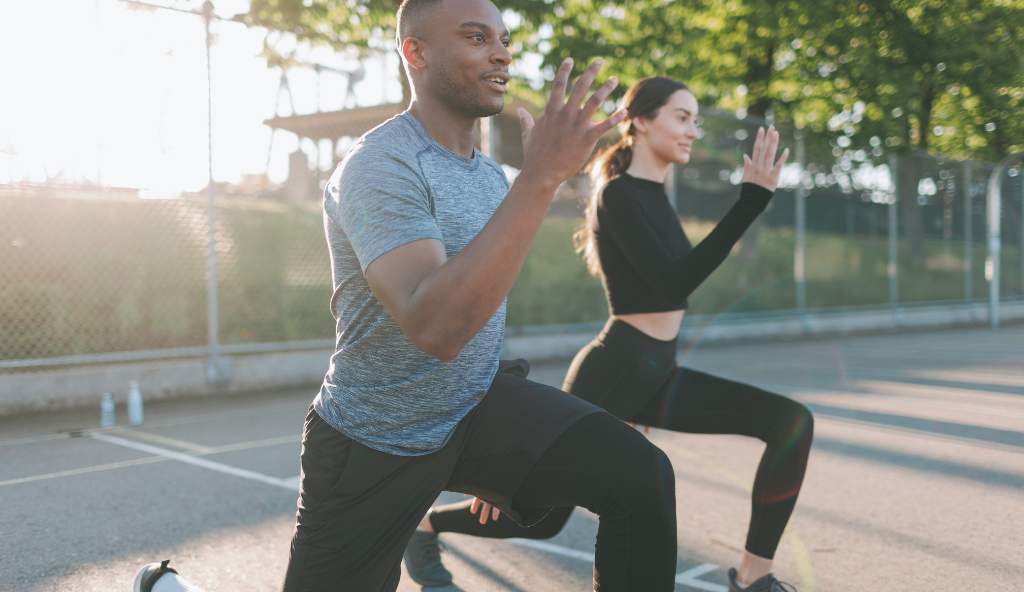 A man and a woman performing lunges outdoors on a sports court, dressed in athletic wear, with a bright sunny background wellness