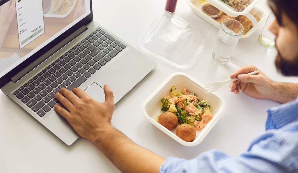 A man in a blue shirt eating a meal from a takeaway container while working on a laptop, with an online food ordering page open on the screen