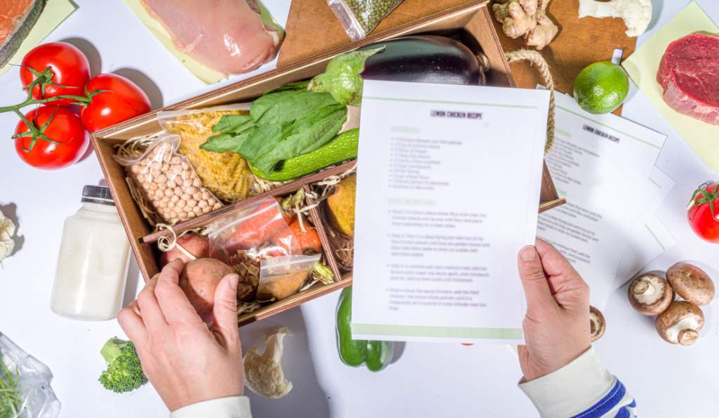 A person holding a printed recipe card while unpacking a meal kit box filled with fresh ingredients such as vegetables, pasta, and legumes, surrounded by various food items on a white table food