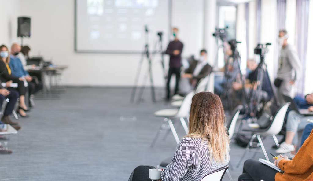 Attendees seated in a modern conference room with cameras set up for recording, a large screen displaying content, and some participants wearing face masks events