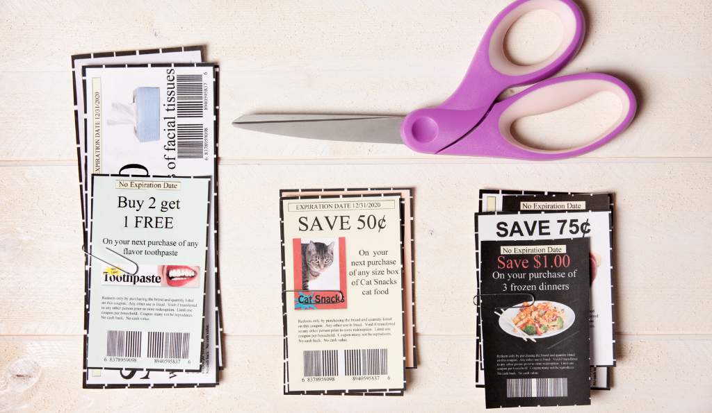 Purple-handled scissors and neatly cut coupons on a light wooden surface, offering discounts on toothpaste, cat snacks, and frozen dinners orinted