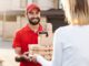 Hungry Howie’s $5, $10, $15 Deals A smiling delivery man in a red uniform and cap handing over pizza boxes and coffee cups to a customer wearing a white sweater outside a residential area food