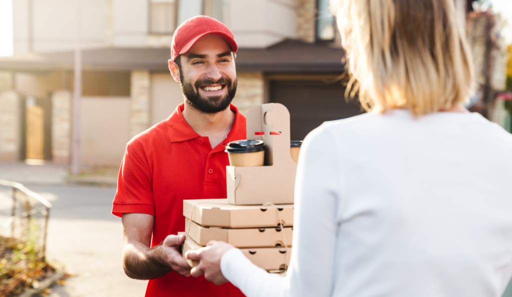 A smiling delivery man in a red uniform and cap handing over pizza boxes and coffee cups to a customer wearing a white sweater outside a residential area food