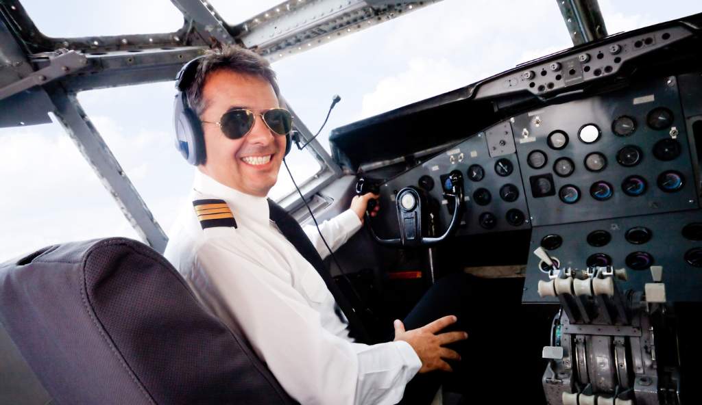 Smiling pilot wearing sunglasses and a headset, sitting in the cockpit of an airplane, holding the control yoke flights