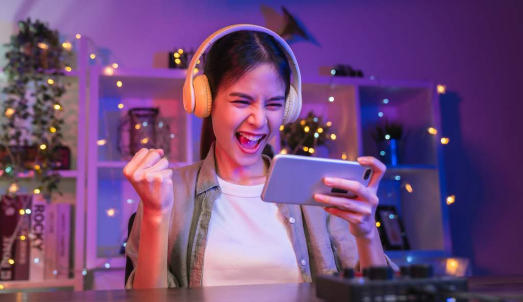 Excited young woman wearing headphones, celebrating while playing a mobile game on her smartphone in a colorful, illuminated gaming setup