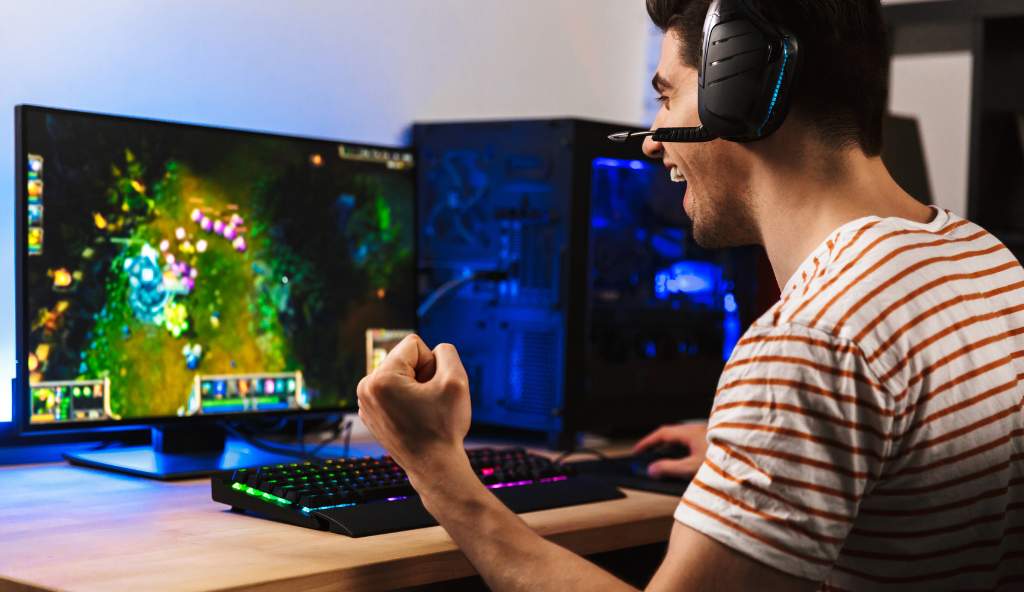 Young man wearing a gaming headset, celebrating while playing a video game on a high-performance PC setup with a large monitor and RGB keyboard