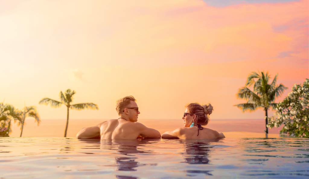 Romantic couple relaxing in an infinity pool at sunset, overlooking the ocean with palm trees in the background hotel