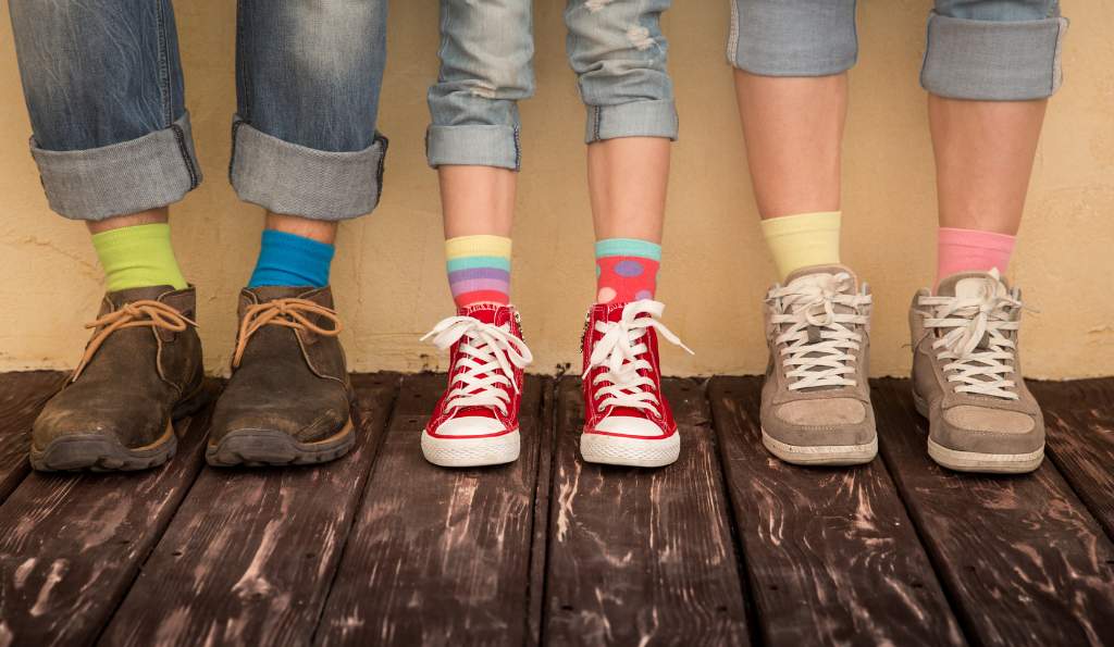 Close-up of four people standing side by side on a wooden floor, wearing rolled-up jeans, colorful mismatched socks, and different types of shoes, including boots, sneakers, and high-tops