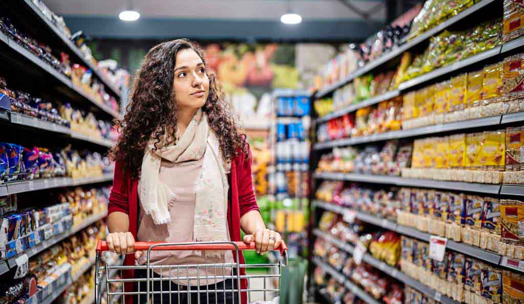 A young woman with curly hair wearing a red cardigan and a scarf, pushing a shopping cart through a supermarket aisle filled with various packaged goods