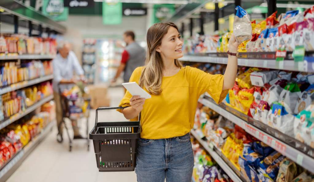 A young woman in a yellow top shopping in a supermarket aisle, holding a shopping basket and a notepad while examining a packaged product