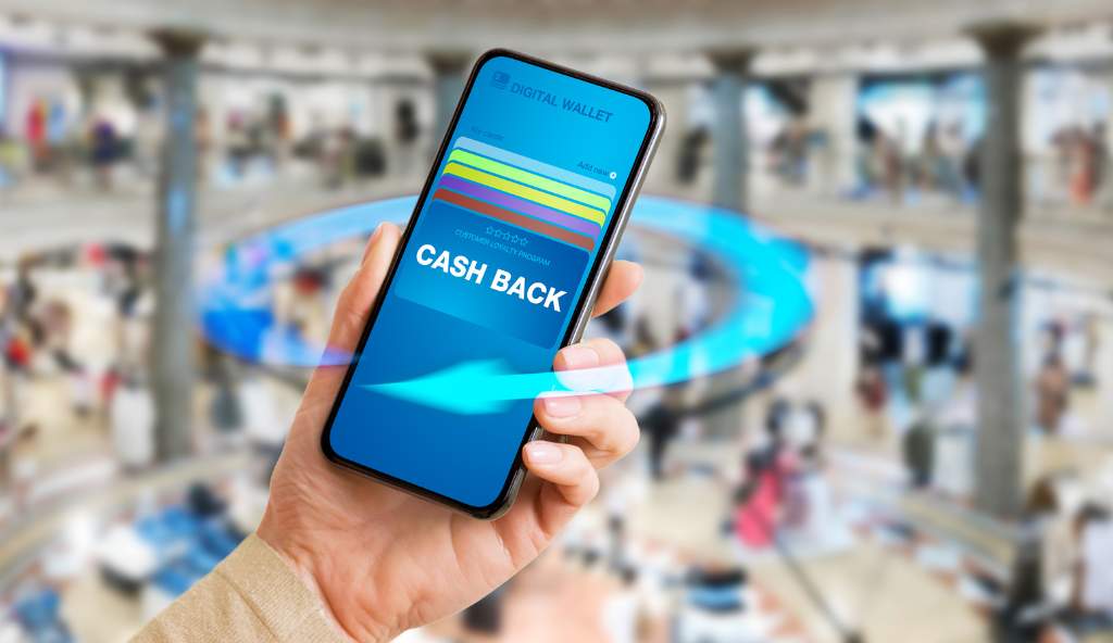 Hand holding a smartphone displaying a digital wallet with a 'CASH BACK' offer, with a blurred shopping mall background, representing digital payments and rewards bundles