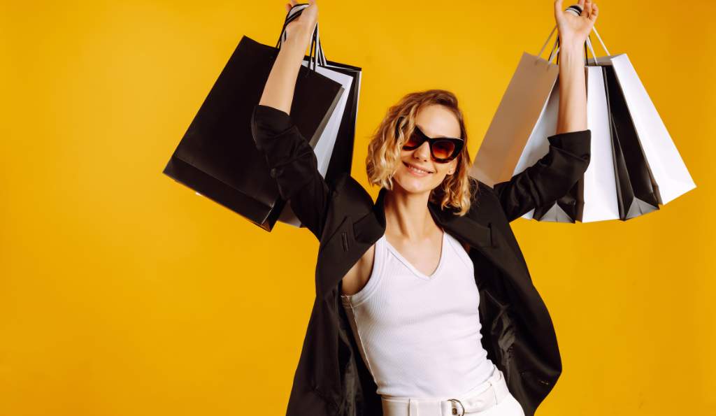 A stylish woman wearing sunglasses, a black blazer, and a white top, holding multiple black and white shopping bags in both hands, smiling against a vibrant yellow background