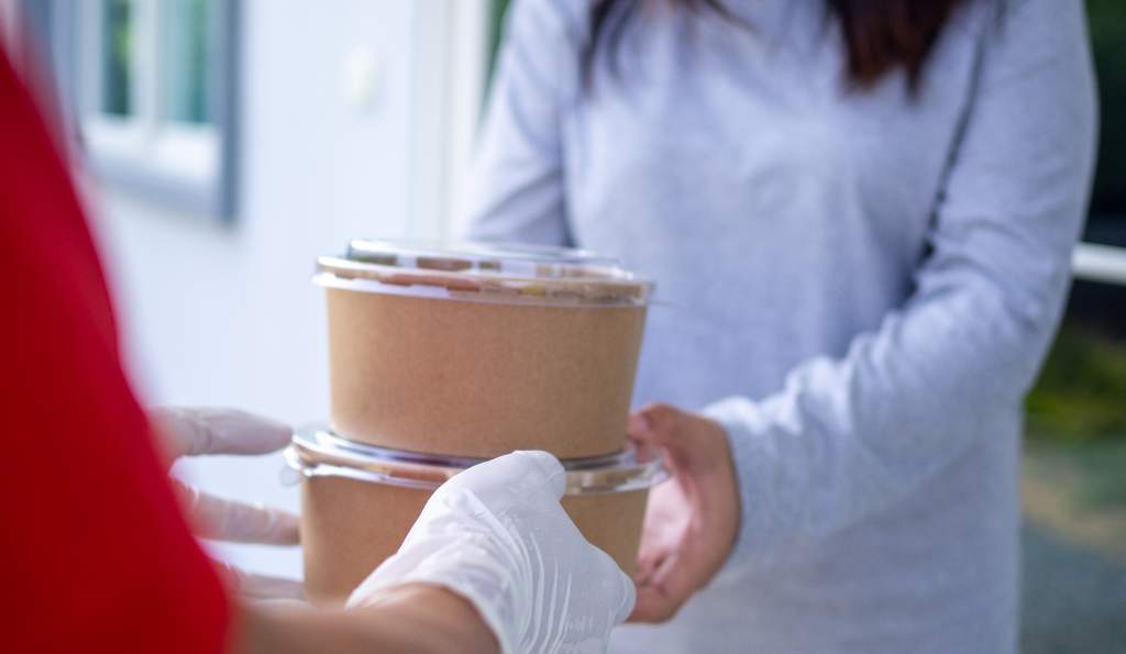 A delivery person wearing gloves hands over two takeaway food containers to a woman in a grey sweater at her doorstep food