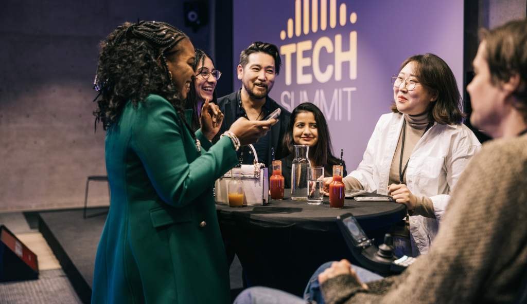 Diverse group of professionals networking and laughing at a tech summit, gathered around a table with drinks and mobile devices events