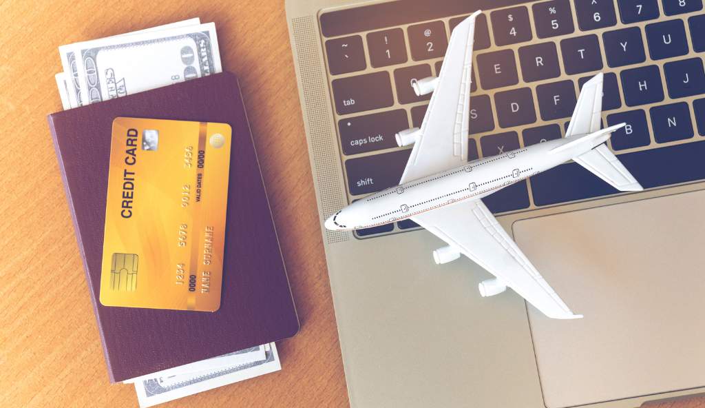 Travel essentials including a passport with cash, a credit card, a laptop keyboard, and a toy airplane symbolizing online flight booking and travel planning