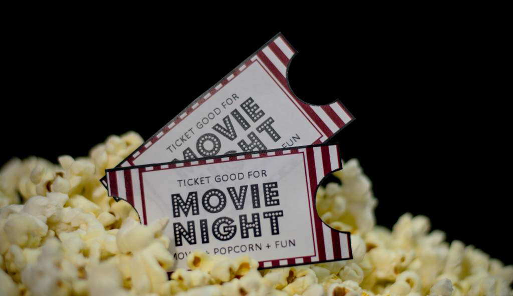 Two movie night tickets with a red and white striped design placed on a pile of popcorn against a black background