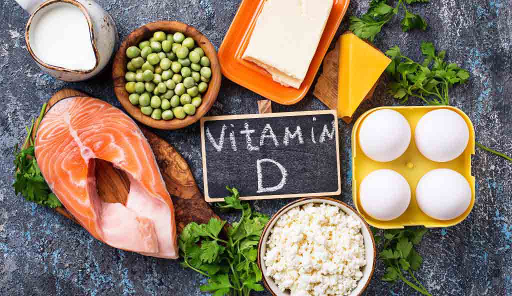 A variety of foods rich in Vitamin D, including salmon, eggs, milk, butter, cheese, green peas, and cottage cheese, arranged around a small chalkboard with 'Vitamin D' written on it