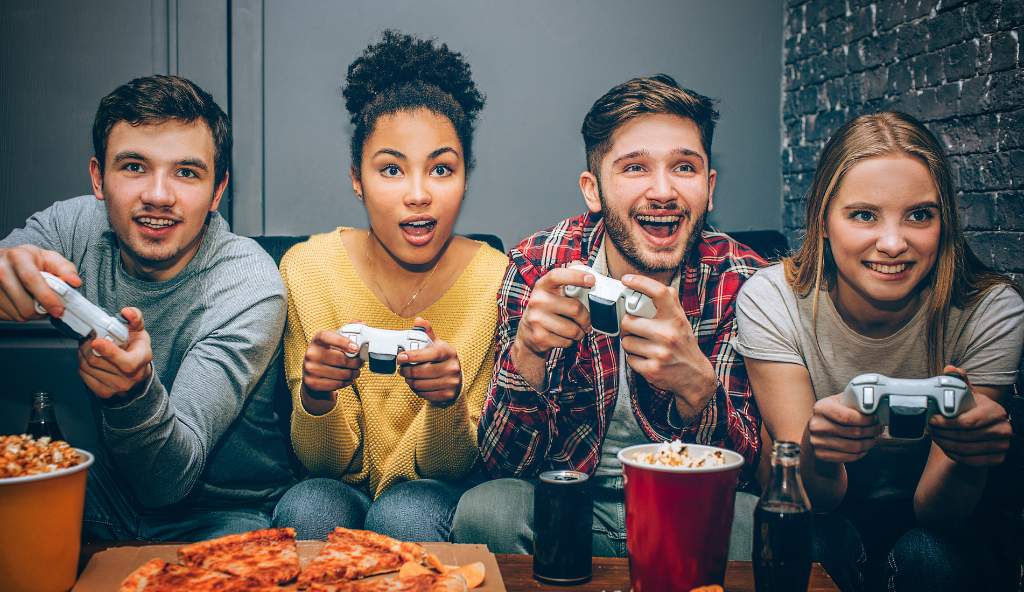 Group of four young friends excitedly playing a video game together with controllers, surrounded by pizza, popcorn, and drinks in a cozy gaming setup