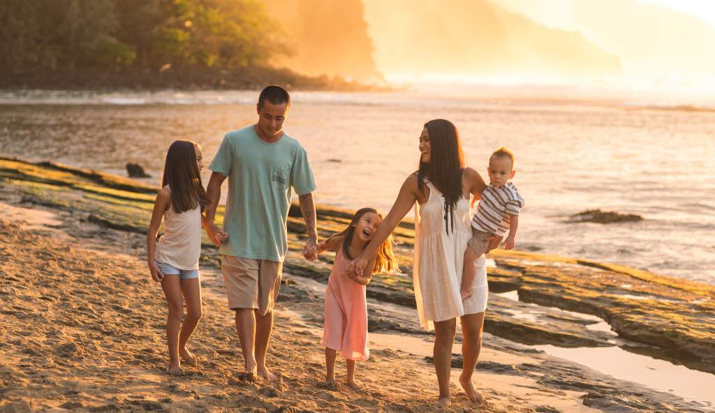 Smiling family of four walking barefoot on a sandy beach at sunset, holding hands and enjoying the warm golden light by the ocean vacation