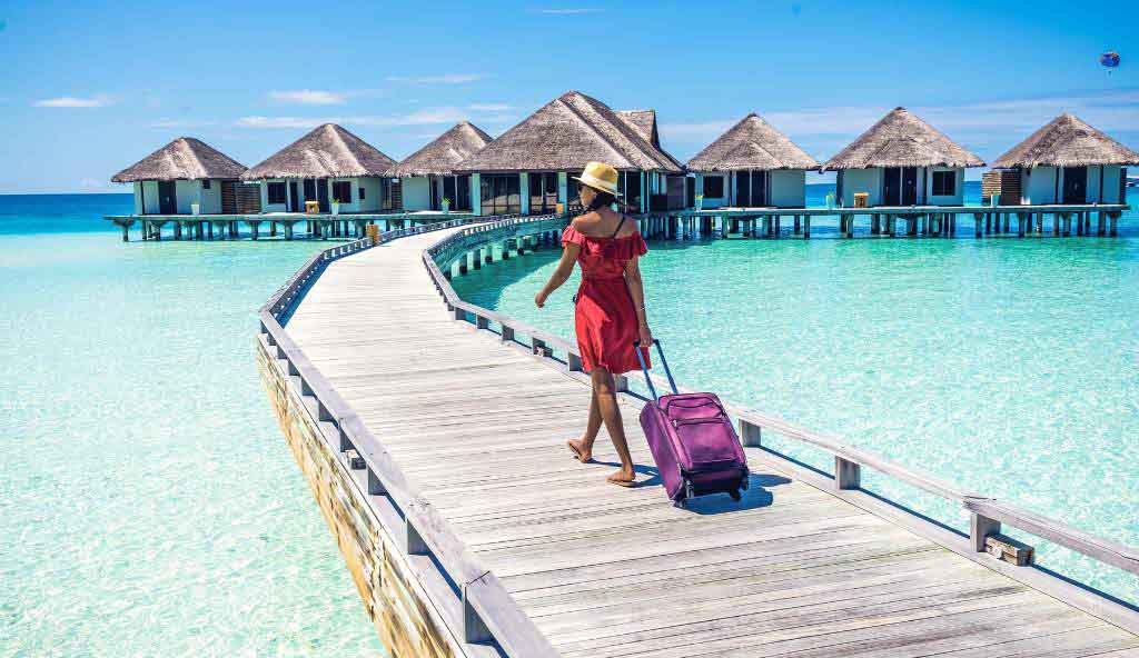 Woman in a red dress and straw hat walking barefoot on a wooden boardwalk over turquoise water, pulling a purple suitcase towards overwater bungalows in a tropical resort vacation