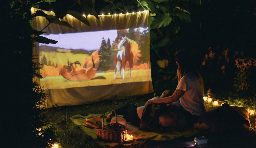 A woman sitting on a blanket in a garden at night, watching an animated movie projected onto a white sheet, surrounded by fairy lights and picnic items