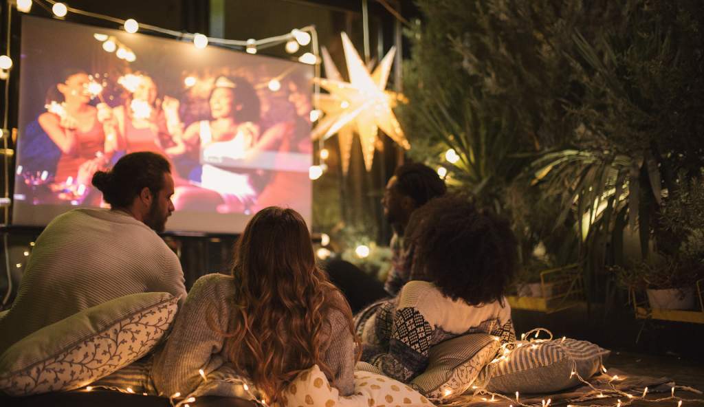 Group of friends watching an outdoor movie screening at night, surrounded by fairy lights and cozy cushions