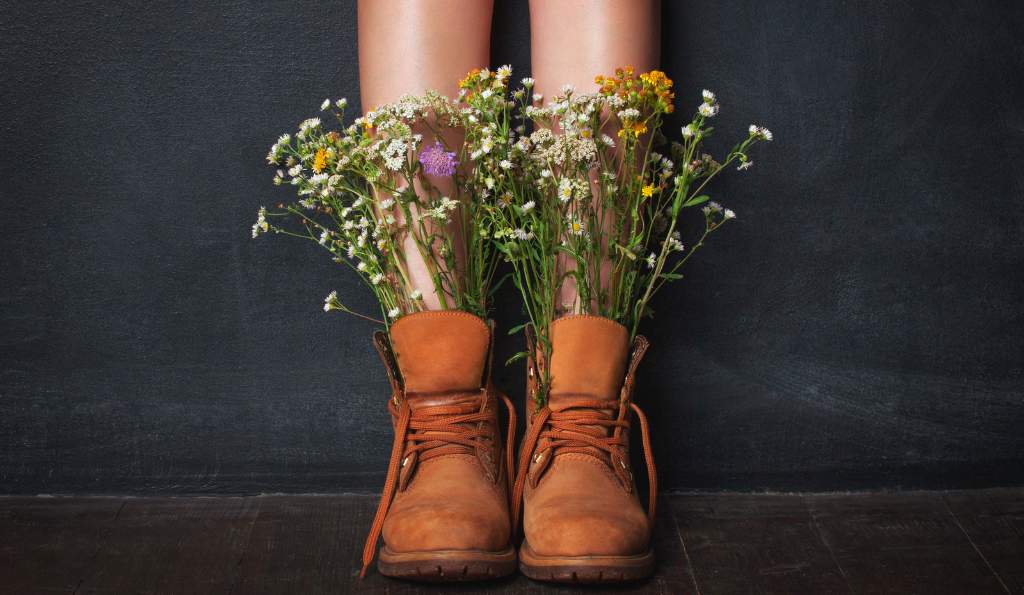 Close-up of a person wearing brown leather boots with wildflowers growing out of them, set against a dark background shoes