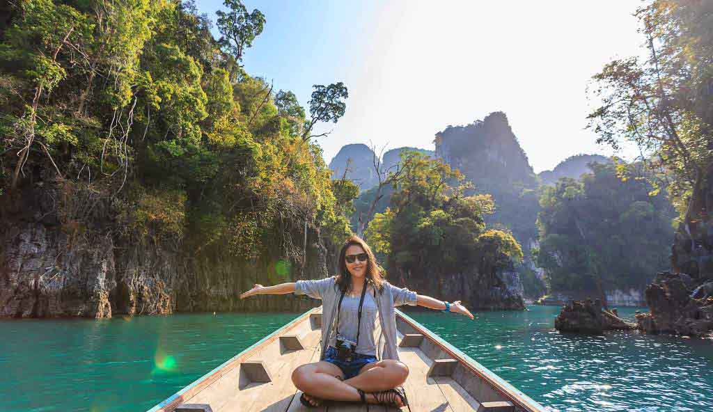 Happy woman sitting cross-legged on the front of a wooden boat, arms outstretched, surrounded by turquoise water and lush green limestone cliffs vacation