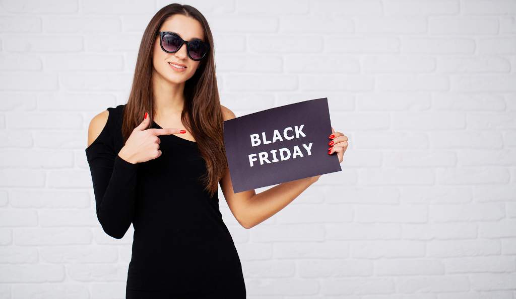 A woman wearing sunglasses and a stylish black dress stands against a white brick wall, smiling and pointing to a black sign that reads 'BLACK FRIDAY' in bold white letters