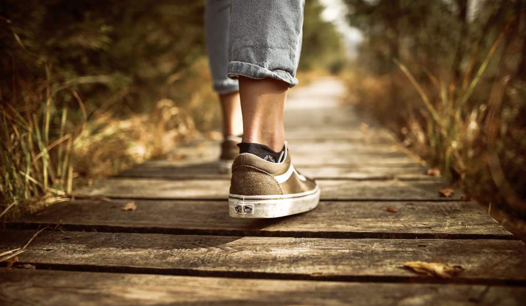 Close-up of a person wearing worn-out sneakers and cropped jeans, walking on a rustic wooden pathway surrounded by nature